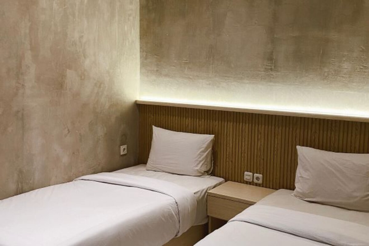 A small, modern hotel room with two single beds, wooden headboard, and a concrete accent wall.