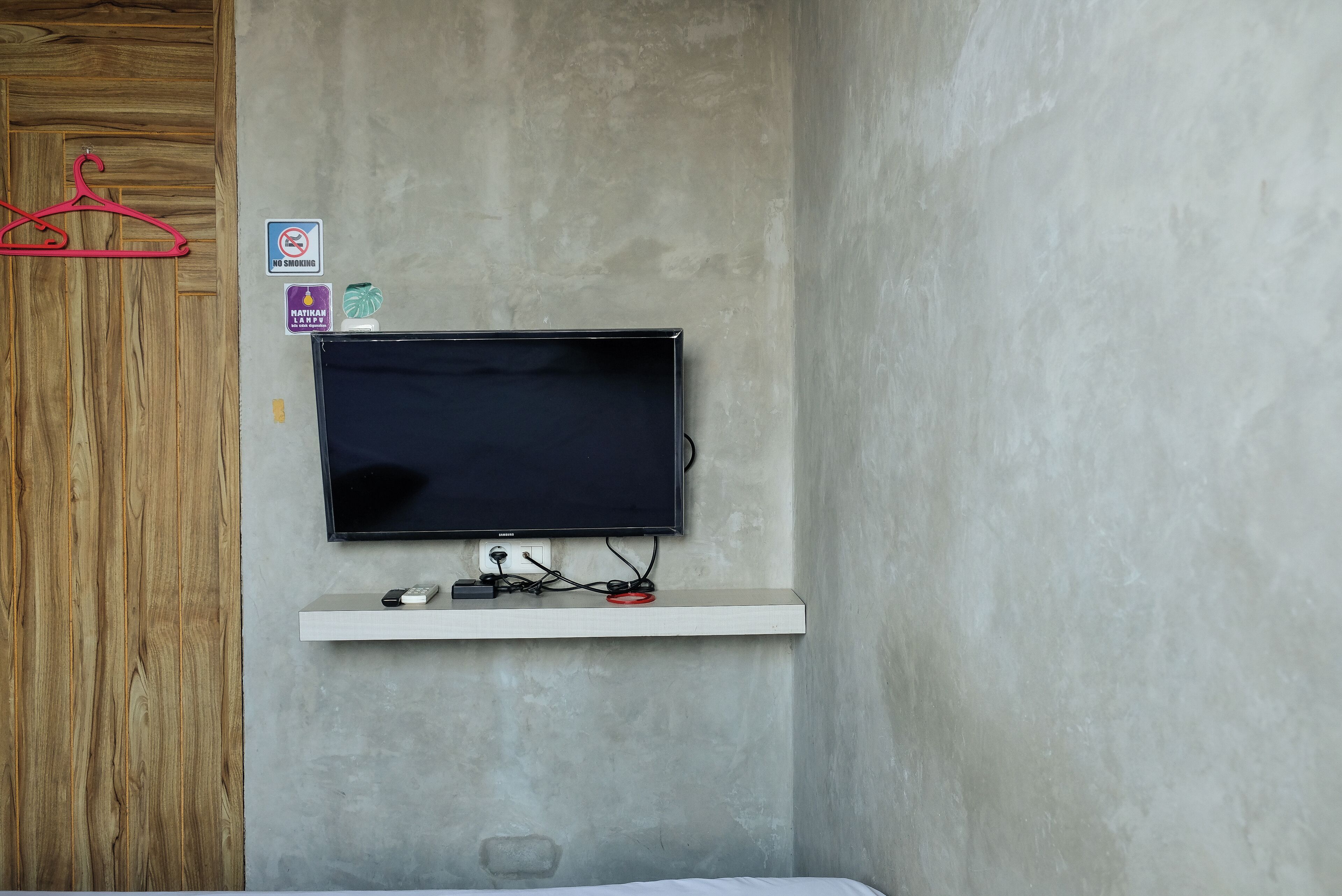 A room with a mounted TV on a white shelf, a wooden door, and a no-smoking sign on the wall.