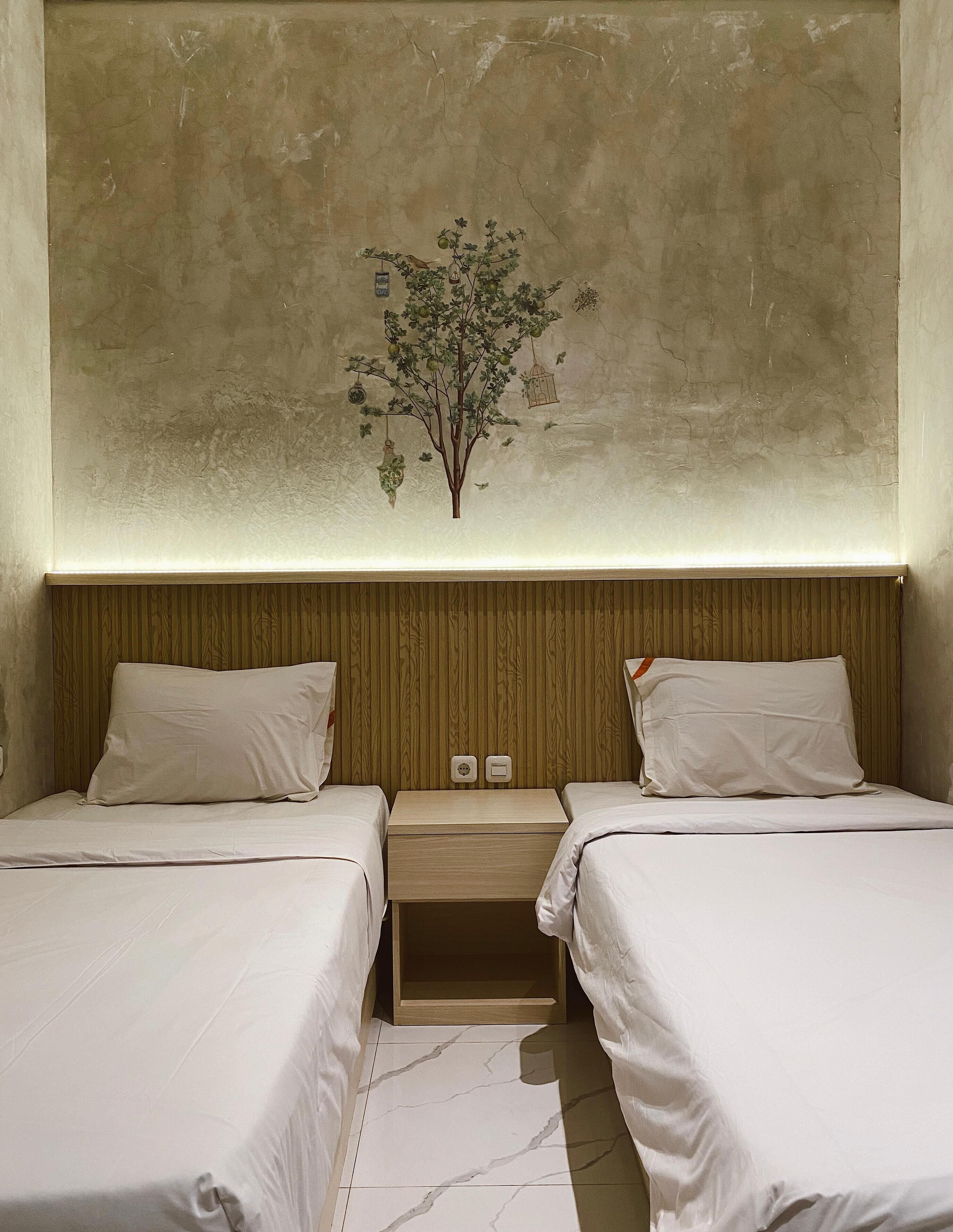 Une chambre d’hôtel avec deux lits, une tête de lit en bois, une table de chevet avec une lampe et une fresque représentant un arbre sur le mur.