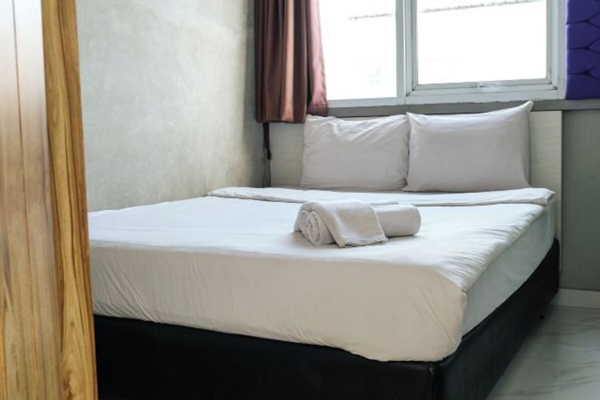 A neatly made bed with white linens and a rolled towel, a window with curtains, and a wooden door frame.