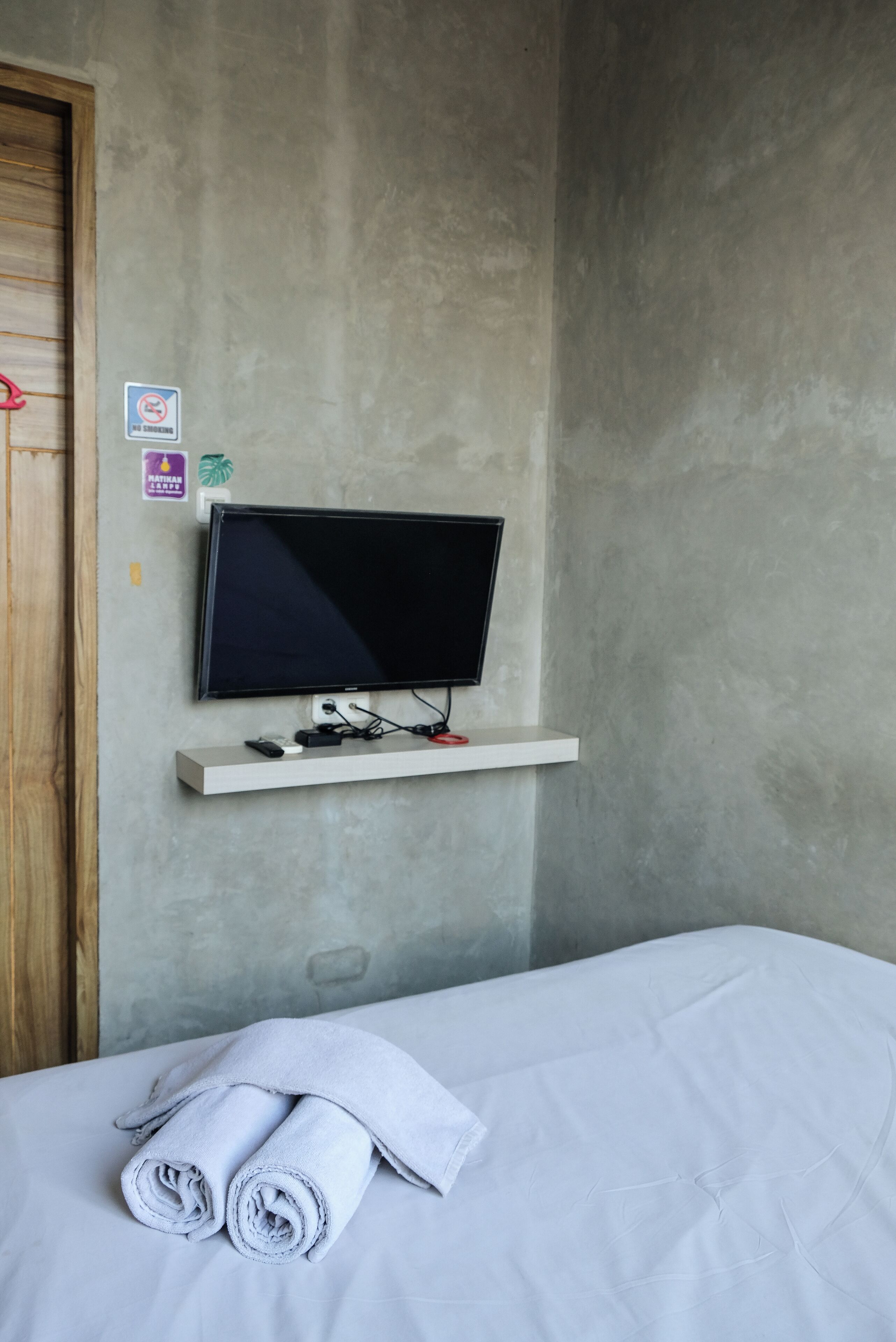 Una habitación con una cama, un televisor montado y un estante en la pared.