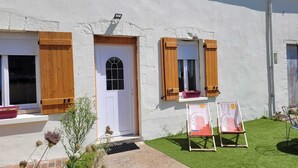 Un bâtiment blanc avec une porte blanche, des volets en bois et une petite fenêtre ornée d’un rideau. On peut voir deux chevalets avec des tableaux posés sur l’herbe, devant le bâtiment.