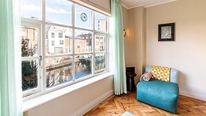 Une pièce avec une grande fenêtre, un canapé bleu, une petite table et un tableau encadré accroché au mur.