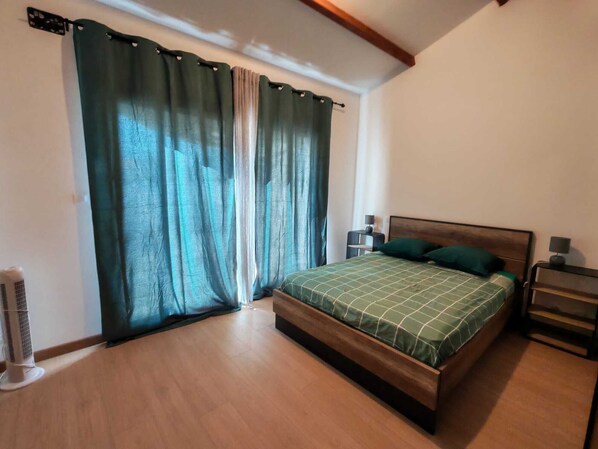 Un dormitorio con cama de madera, cortinas verdes, una mesita de noche y un ventilador de pie.