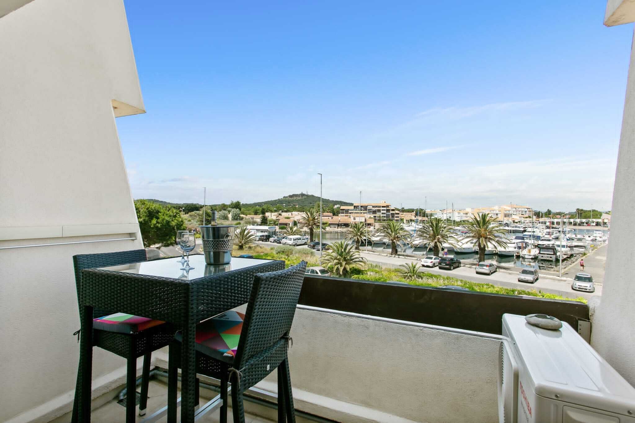 A balcony with a table and chairs, overlooking a marina with boats and a hilly landscape in the distance.