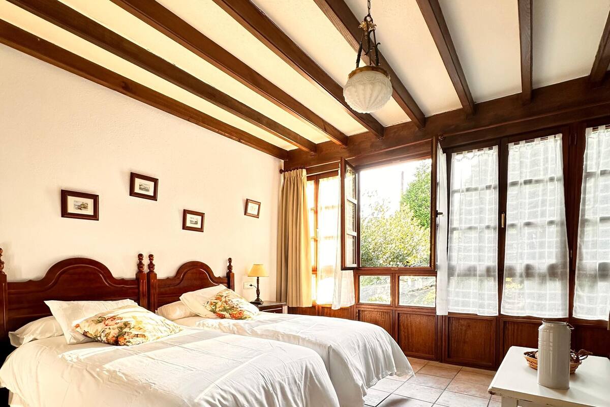 Un dormitorio con una cama doble, cabecera de madera, mesita de noche, lámpara, ventana con cortinas y suelo de baldosas.