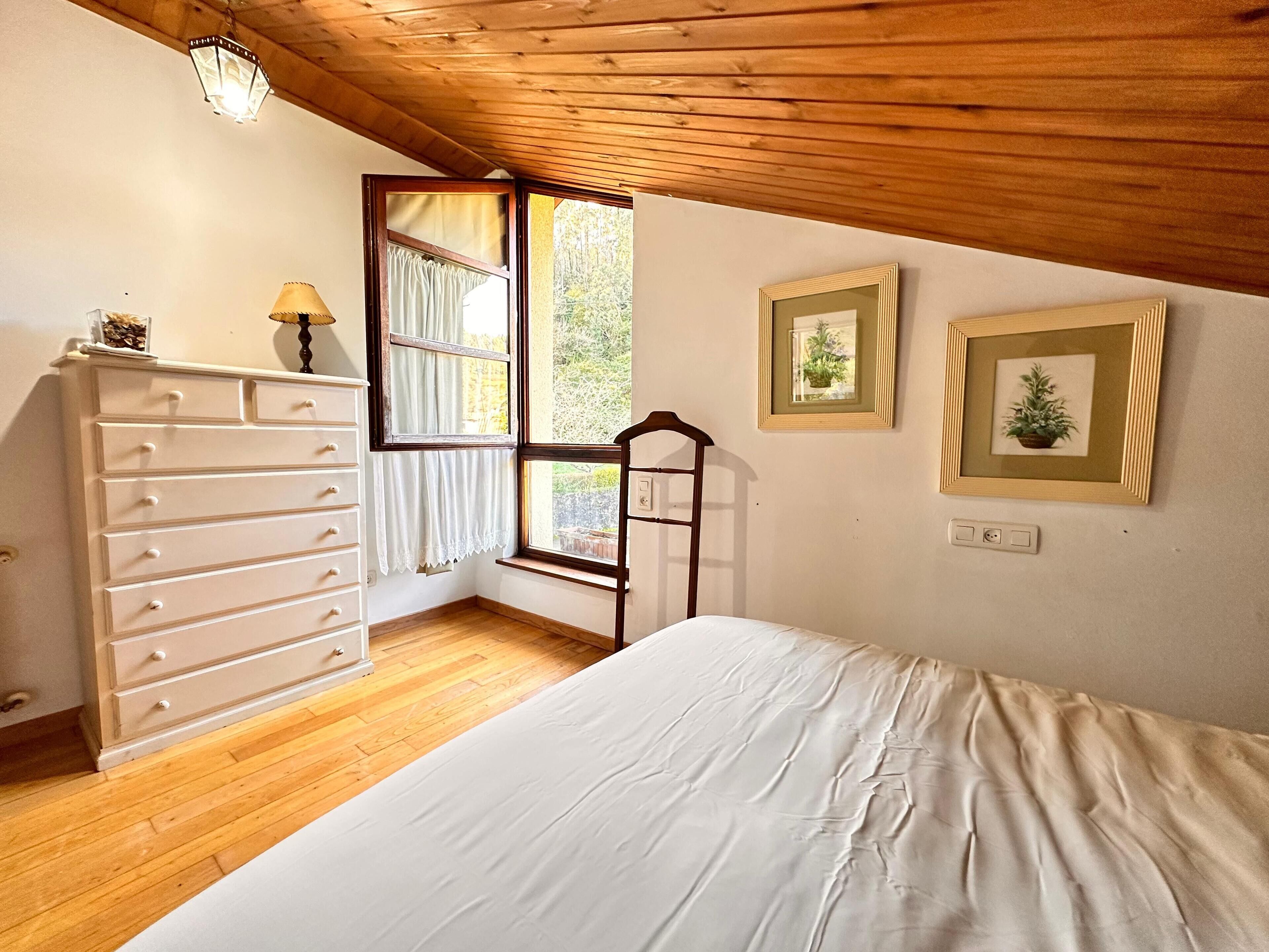 Um quarto com teto de madeira, uma cama, cômoda, janela com cortinas e quadros nas paredes.