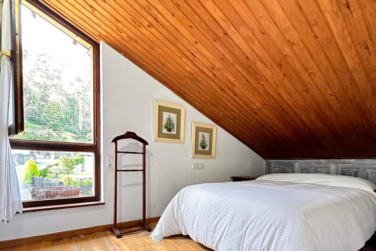 Un dormitorio con techo inclinado, piso de madera, una cama, un ventanal con vistas a la vegetación y dos cuadros en la pared.