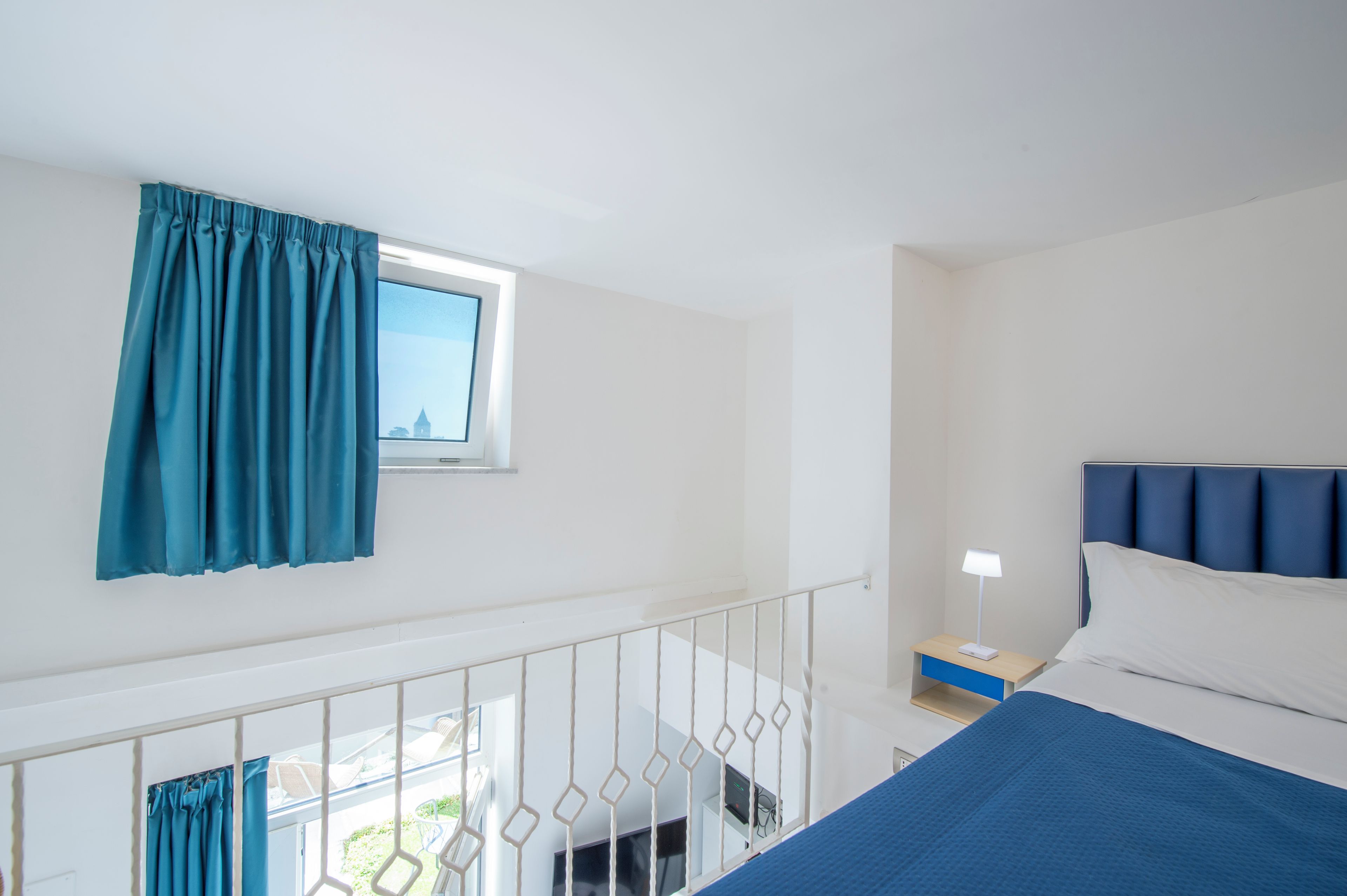 Une chambre d’hôtel avec un lit, une table de chevet, une fenêtre avec des rideaux et un balcon doté d’une balustrade.