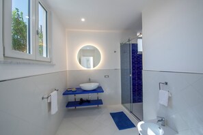 A modern bathroom with a white sink, blue shelves, a round mirror with a light, and a glass shower enclosure. - YourHome - Casa Lucente (Positano)