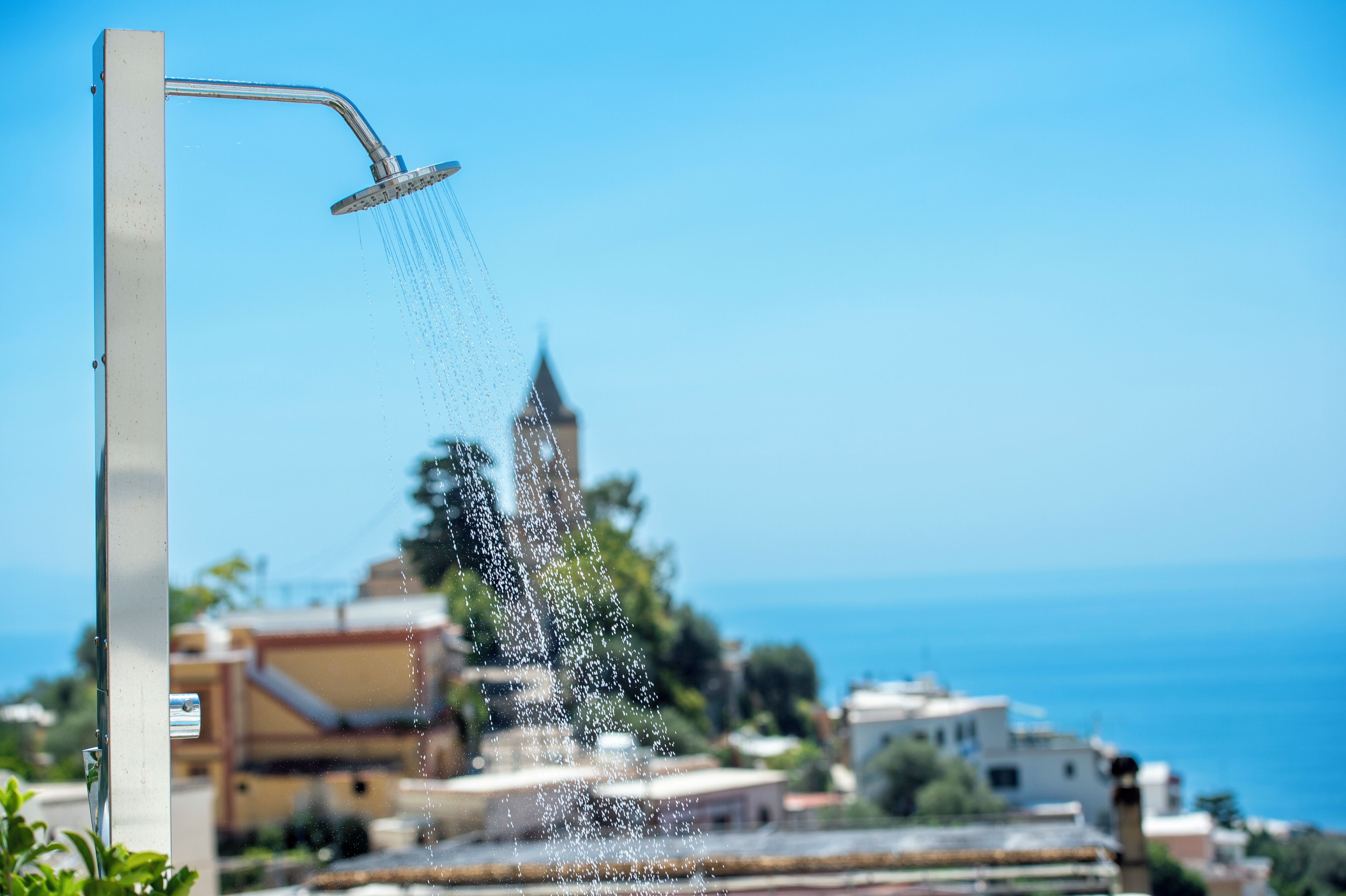 A showerhead with water flowing into the air, a clear blue sky, and a distant view of buildings and a body of water.