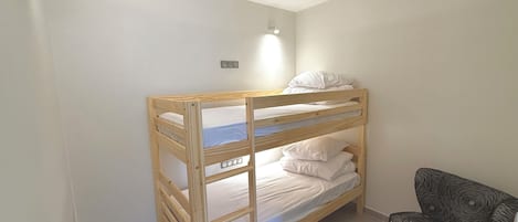 Una cama litera con estructura de madera y ropa de cama blanca, una silla pequeña con estampado negro y un interruptor de luz en la pared.