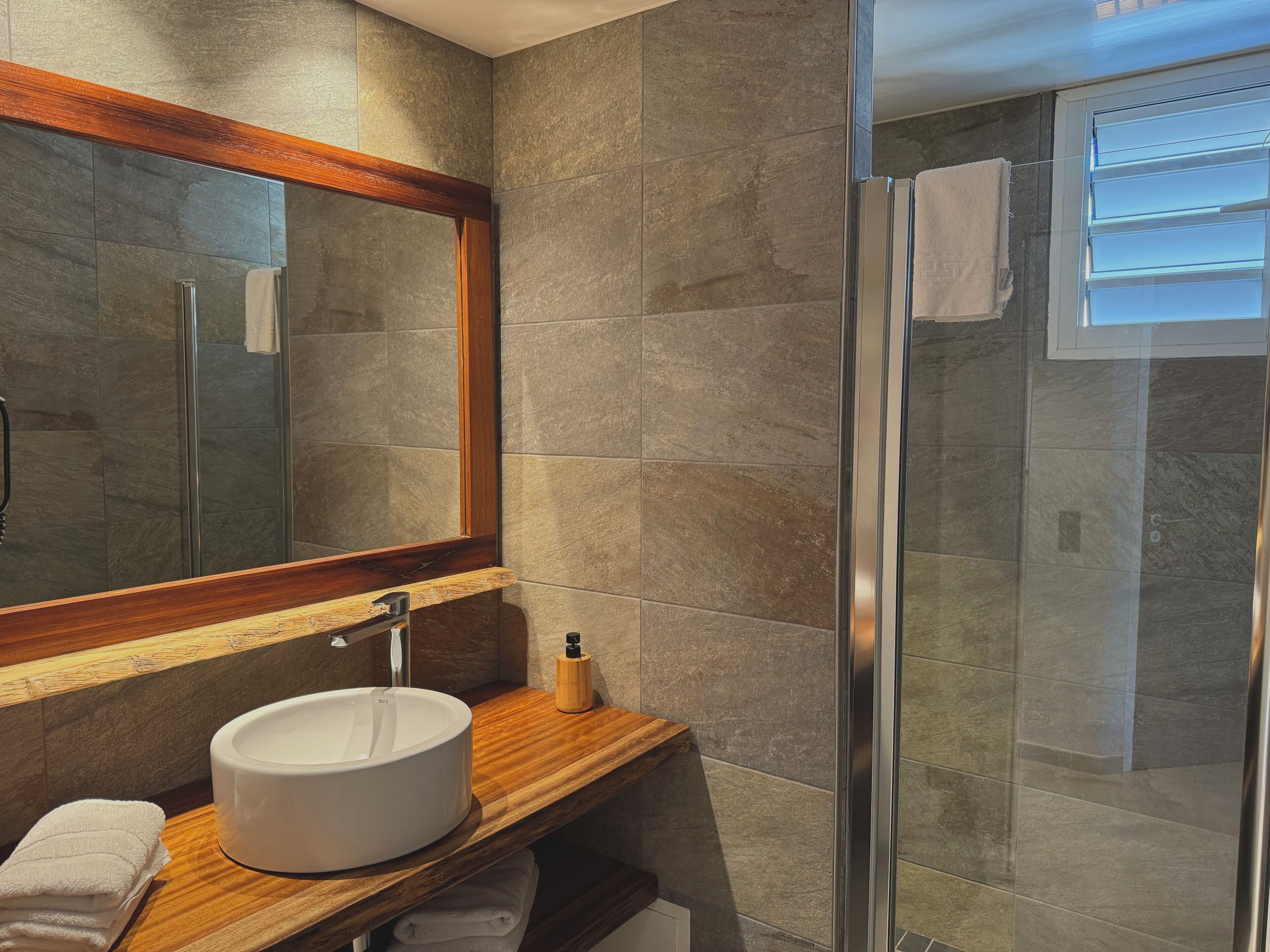 Un baño moderno con un espejo grande, un mueble de tocador de madera, un lavabo redondo y una cabina de ducha de vidrio.