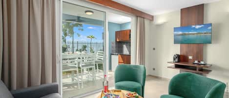 A modern living room with a sofa, a green armchair, and a wooden coffee table with snacks. A TV is mounted on the wall, and a balcony with outdoor furniture is visible through the glass door.