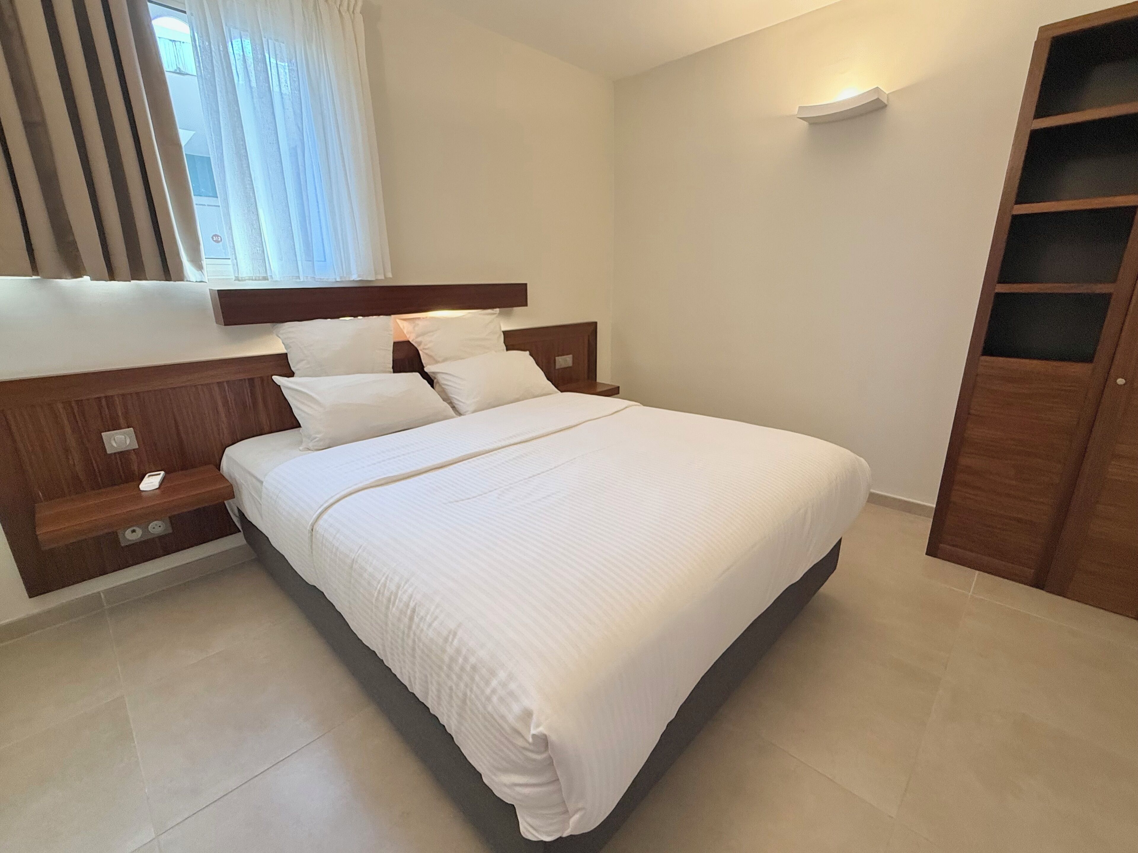 Cama bem arrumada com lençóis brancos em quarto moderno, com cabeceira de madeira, mesa de cabeceira e guarda-roupa.