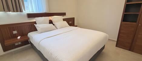 Una cama bien hecha con sábanas blancas en una habitación moderna con cabecera de madera, mesita de noche y armario.