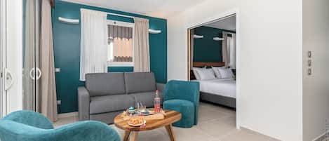 Una habitación moderna con paredes verde azulado, un sofá gris, una mesa de centro de madera con aperitivos, sillones verde azulado y una cama con ropa de cama blanca.