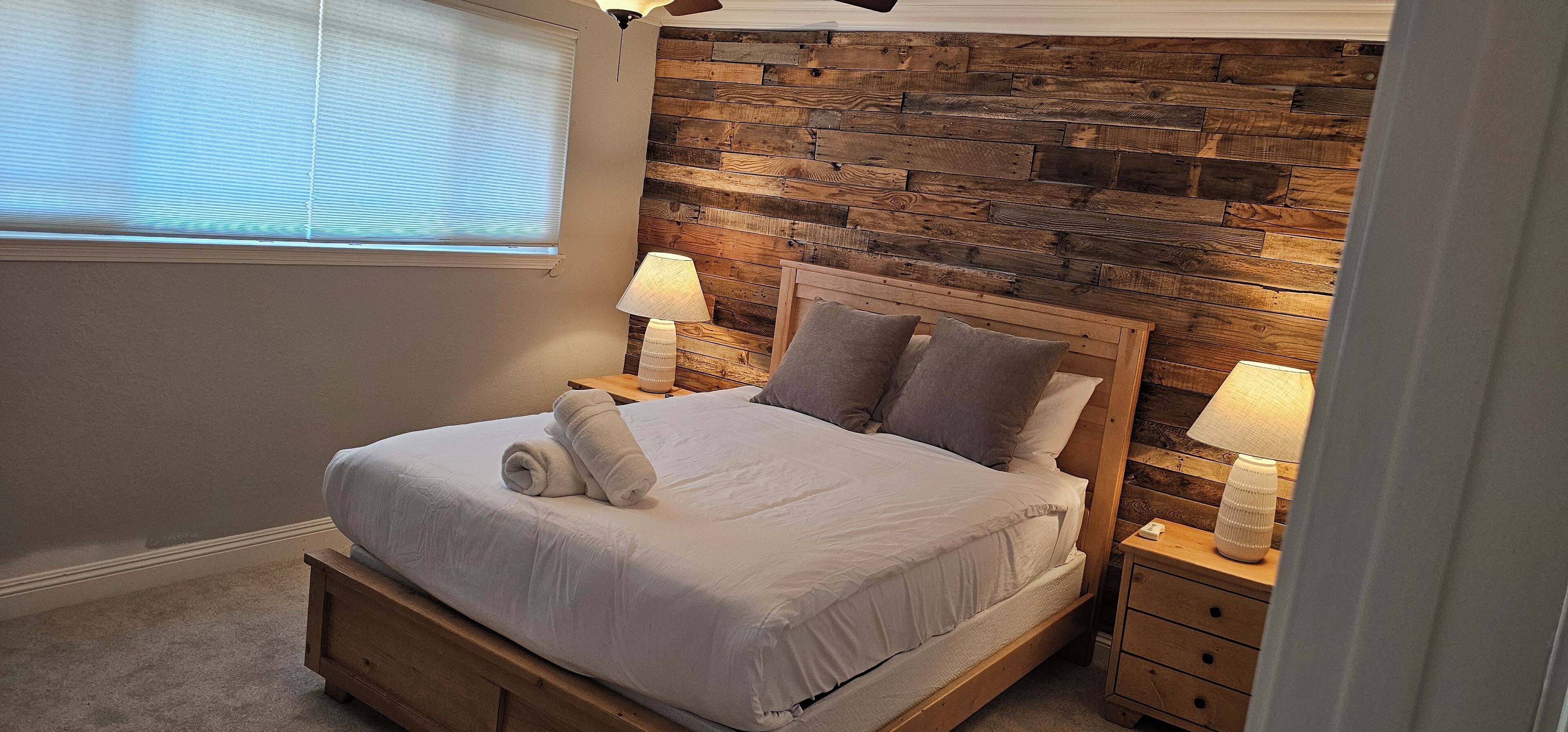Ein Schlafzimmer mit hölzernem Kopfteil, einem Bett mit weißer Bettwäsche, zwei Nachttischlampen und einem Fenster mit Jalousien.