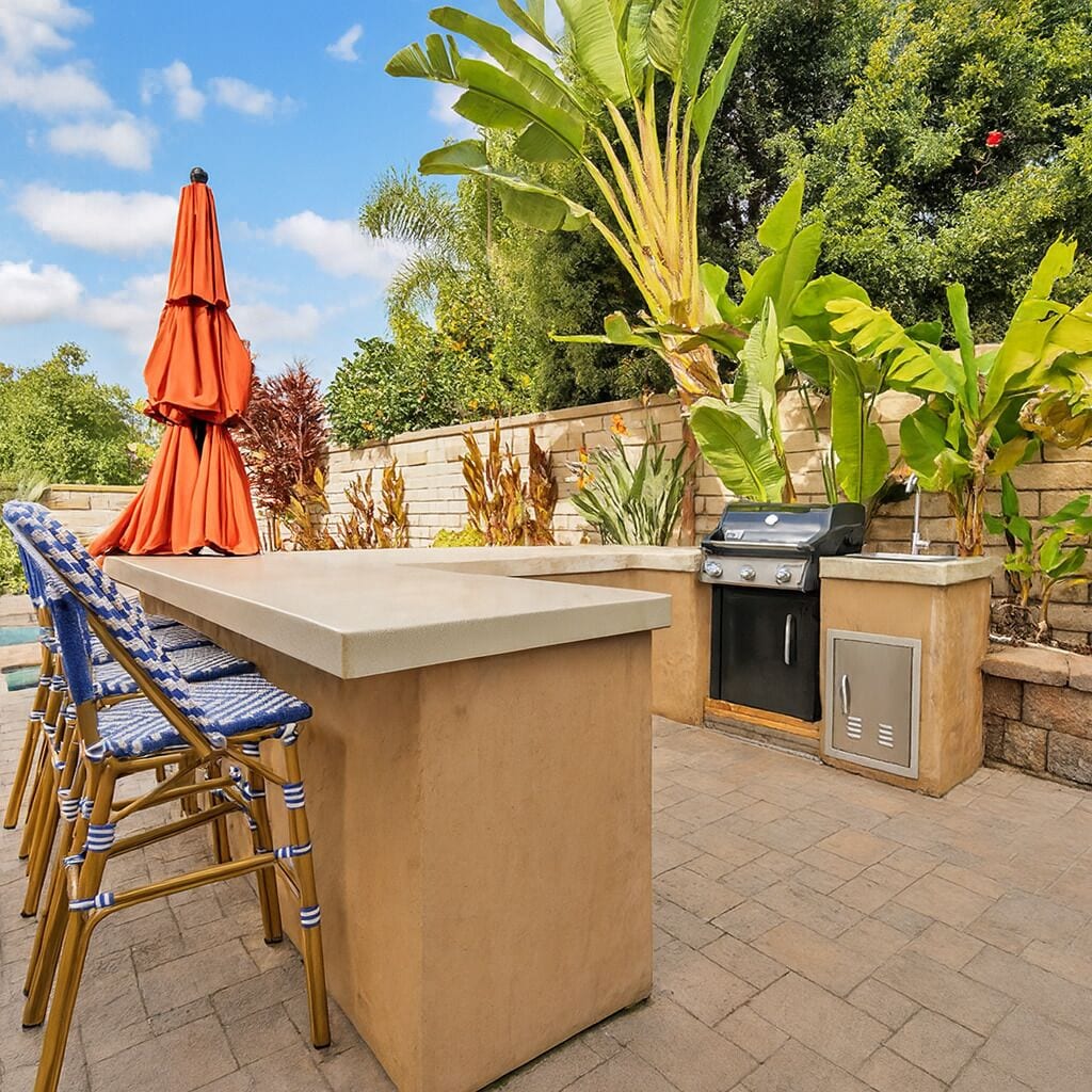 An outdoor kitchen with a grill, bar stools, and a large umbrella.