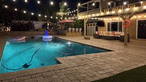 Une piscine équipée d’une pompe à eau, entourée de carreaux de pierre. Des guirlandes lumineuses sont suspendues au-dessus, et une bouée de sauvetage est accrochée à un poteau.