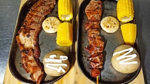 Zwei Tabletts mit gegrilltem Fleisch, Mais und Kartoffeln mit weißer Soße.