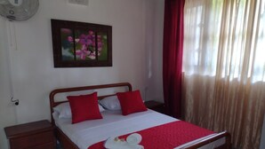 Ein ordentlich bezogenes Bett mit roter und weißer Bettwäsche, ein hölzerner Kopfteil, ein Nachttisch mit Schublade, ein an der Wand befestigter Ventilator und ein gerahmtes Blumenbild über dem Bett.
