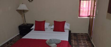 Quarto de hotel com cama, mesa de cabeceira, luminária e janela com cortinas.
