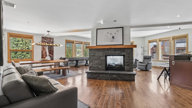 A spacious living room with a stone fireplace, wooden flooring, a gray sofa, and a dining area with a view of the forest. - Rundle Cliffs Lodge by Canmore Premier  (Canmore)