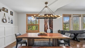 A dining area with a large wooden table, chairs, a chandelier, and a view of mountains through large windows. - Rundle Cliffs Lodge by Canmore Premier  (Canmore)
