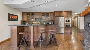 A modern kitchen with wooden cabinets, a central island with stools, stainless steel appliances, and a stone fireplace. - Rundle Cliffs Lodge by Canmore Premier  (Canmore)