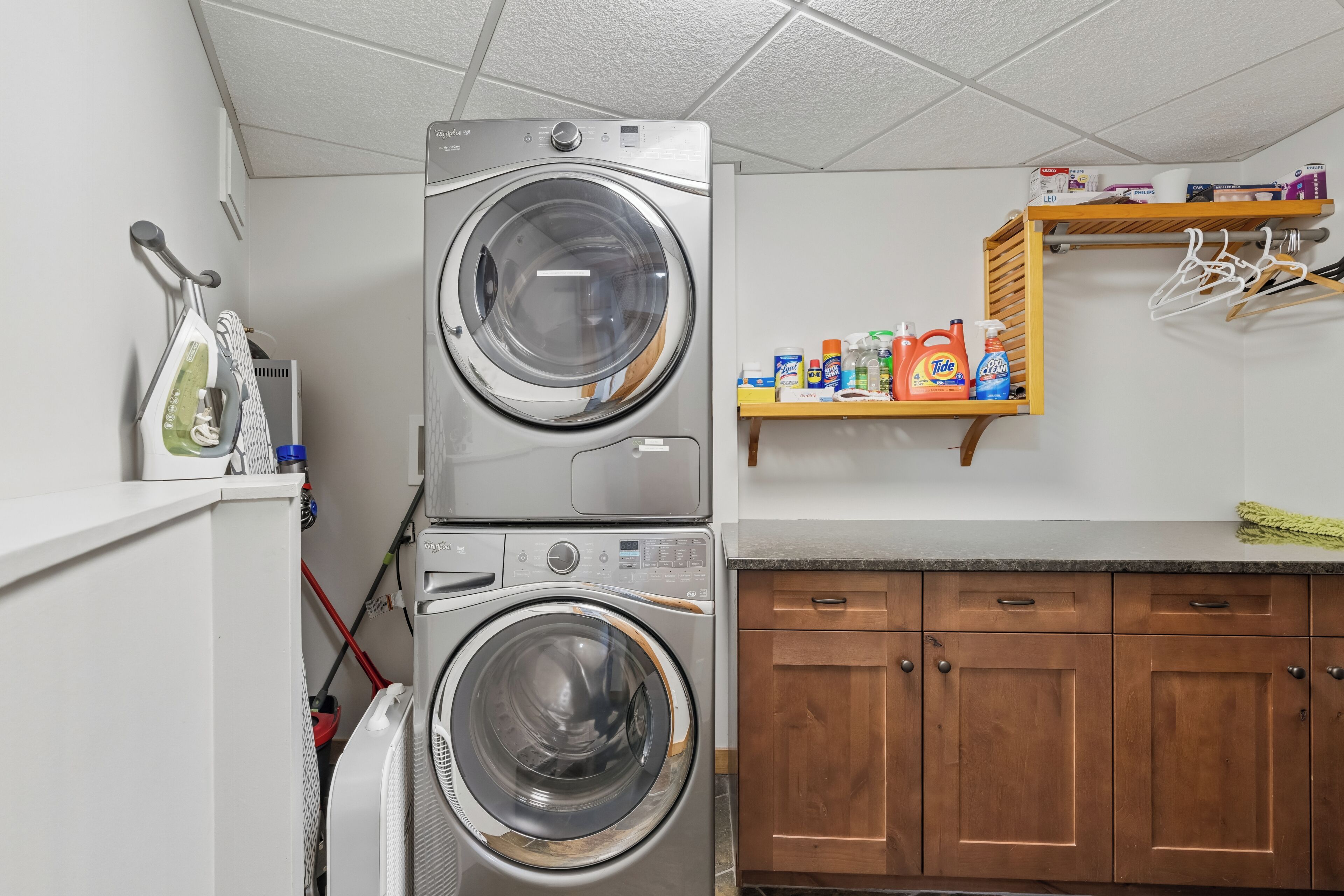 A laundry area with stacked washers and dryers, wooden cabinets, and a shelf with cleaning supplies.