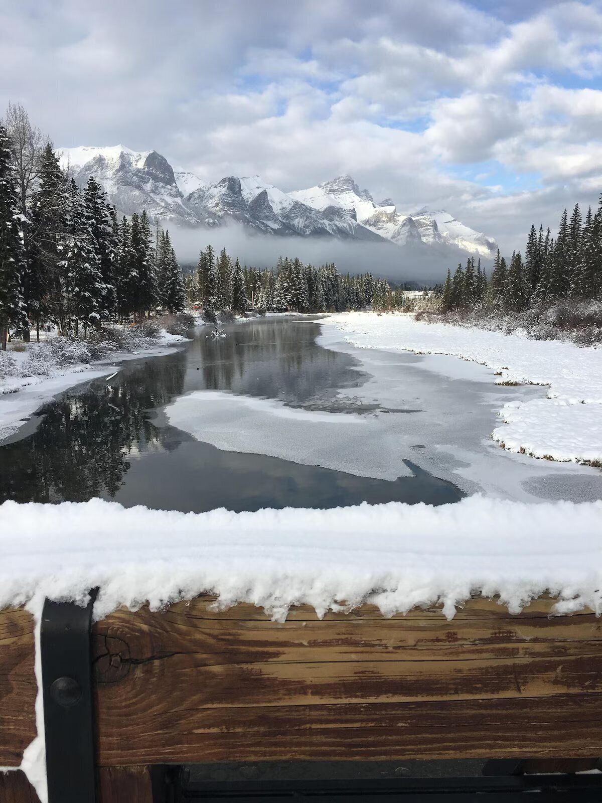 A river with snow-covered banks and trees, surrounded by mountains.
