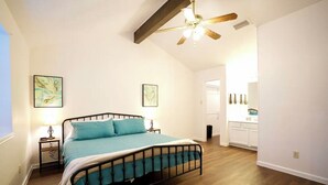 Un dormitorio con una cama, mesitas de noche, ventilador de techo y vistas a la cocina contigua.