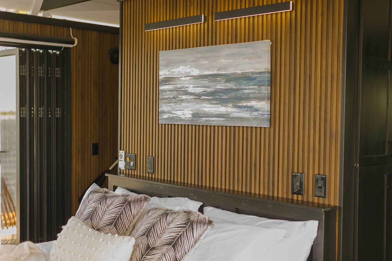 A hotel room with a wooden paneled wall, a bed with patterned pillows, and a painting of a seascape.
