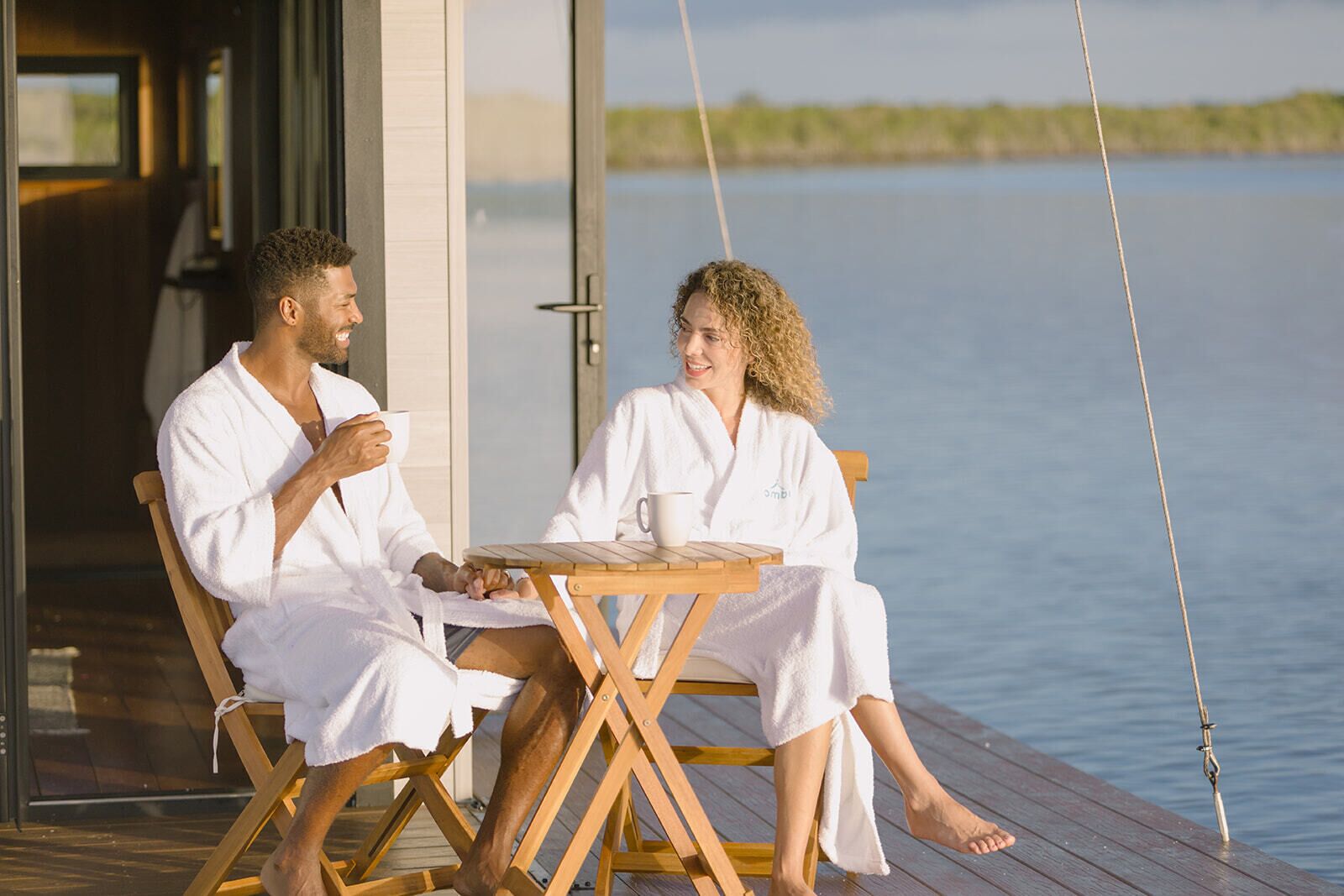 Two people in white bathrobes sitting at a wooden table on a deck overlooking a body of water.