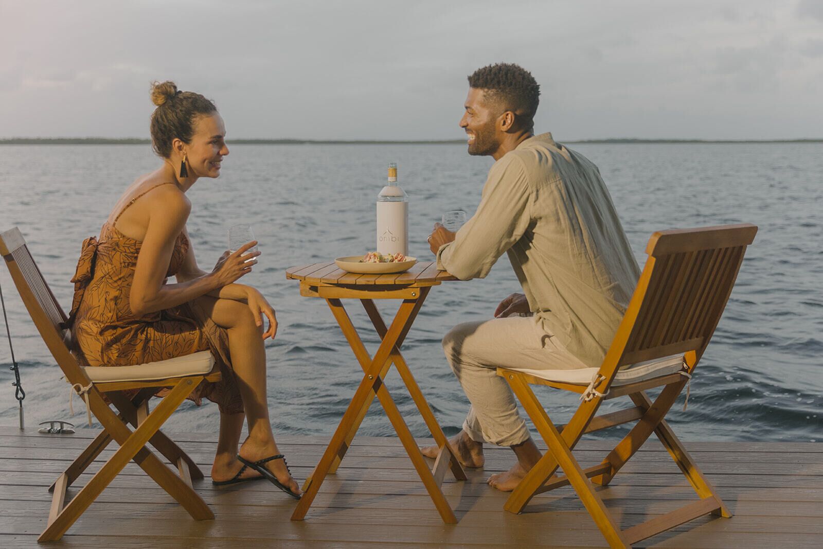 Two people seated at a wooden table on a dock with a view of the water.