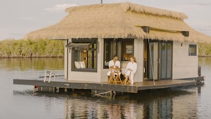 A floating house with a thatched roof, a glass door, and a small deck area. - Premium Floating Bungalow by Stay Ombi (Key Largo)