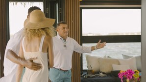 A couple in a room with a large window overlooking the sea, a bed with pillows, a small table with a vase of flowers, and a plant. - Premium Floating Bungalow by Stay Ombi (Key Largo)