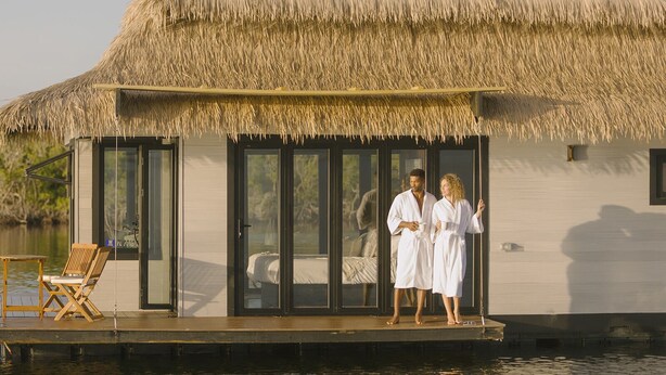 A floating house with a thatched roof, large sliding glass doors, and a wooden deck with a chair. - Premium Floating Bungalow by Stay Ombi (Key Largo)