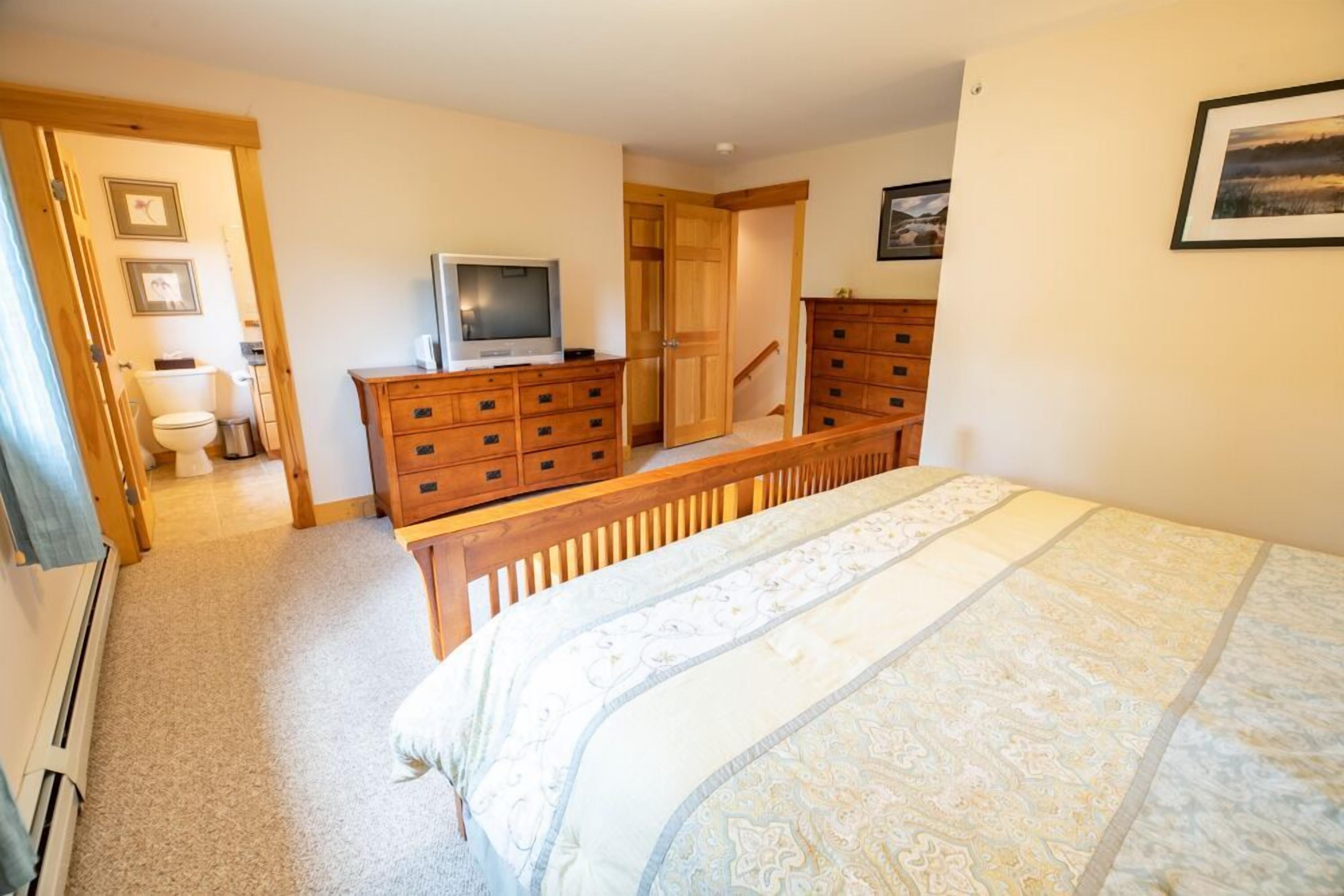Un dormitorio con cama, cómoda de madera, televisor y puerta que da a un baño.