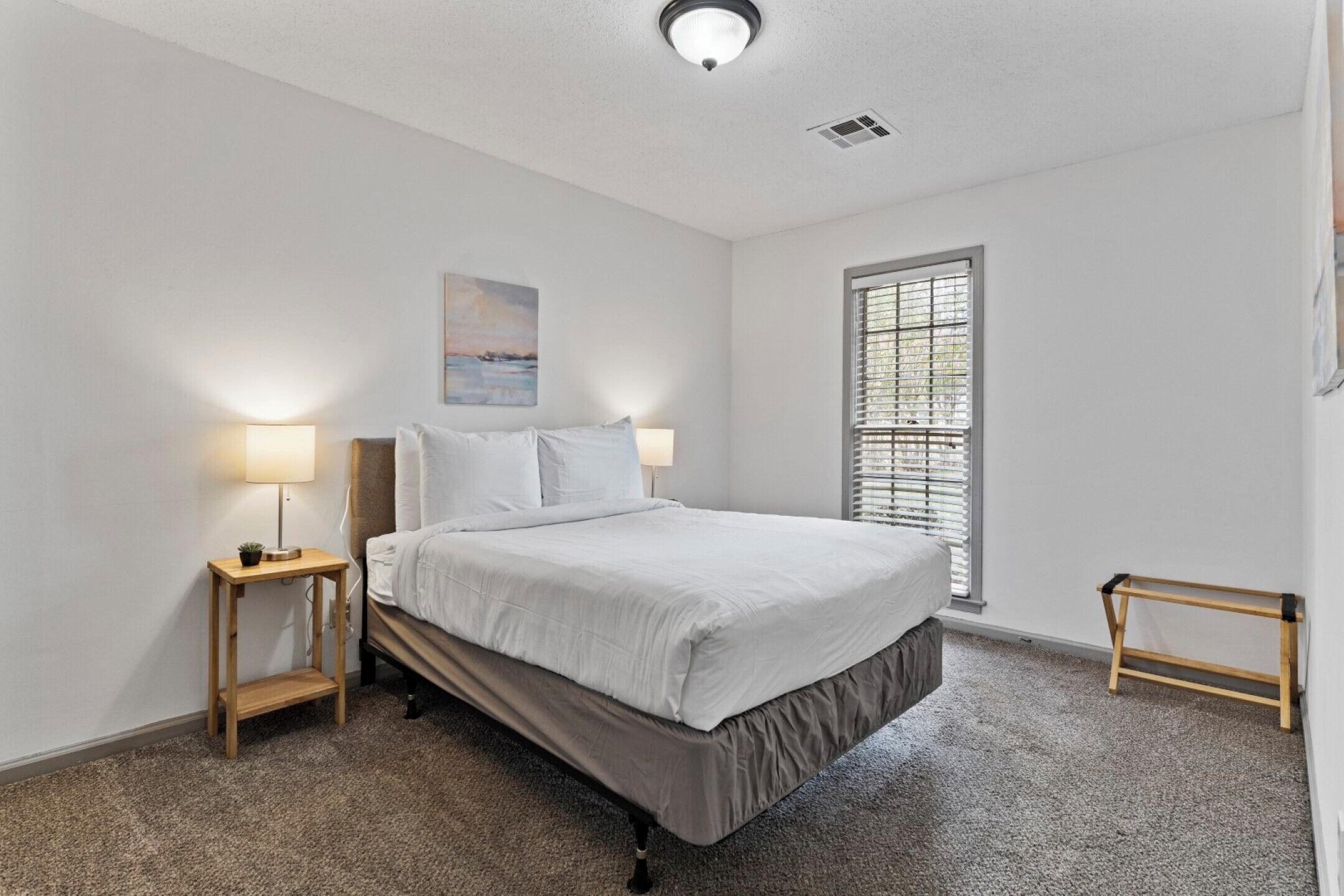 A bedroom with a bed, two bedside tables with lamps, a window with blinds, and a painting on the wall.