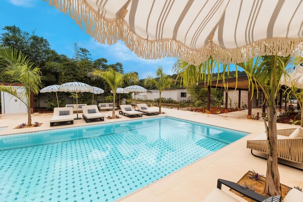 A swimming pool with lounge chairs and umbrellas, surrounded by tropical plants and a wooden structure. - The Banks Resort (Belmopan)