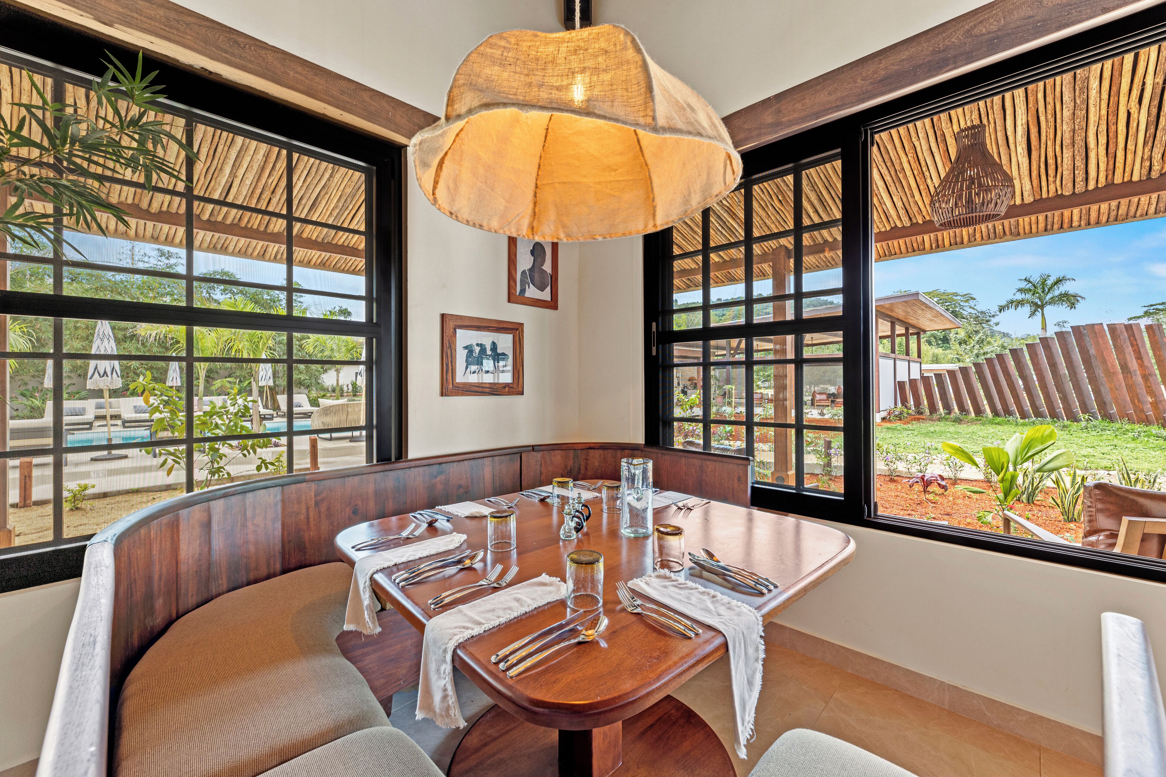 A dining area with a wooden table set for a meal, large windows with a view of the outdoors, and a hanging light fixture.