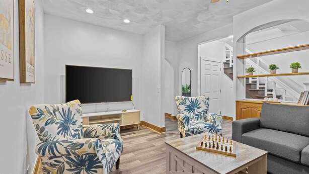 A modern living room with a flat-screen TV, two floral armchairs, a grey sofa, and a coffee table with a chess set.
