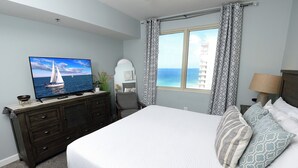 A bedroom with a bed, a TV on a dresser, a window with curtains, and a view of the sea.