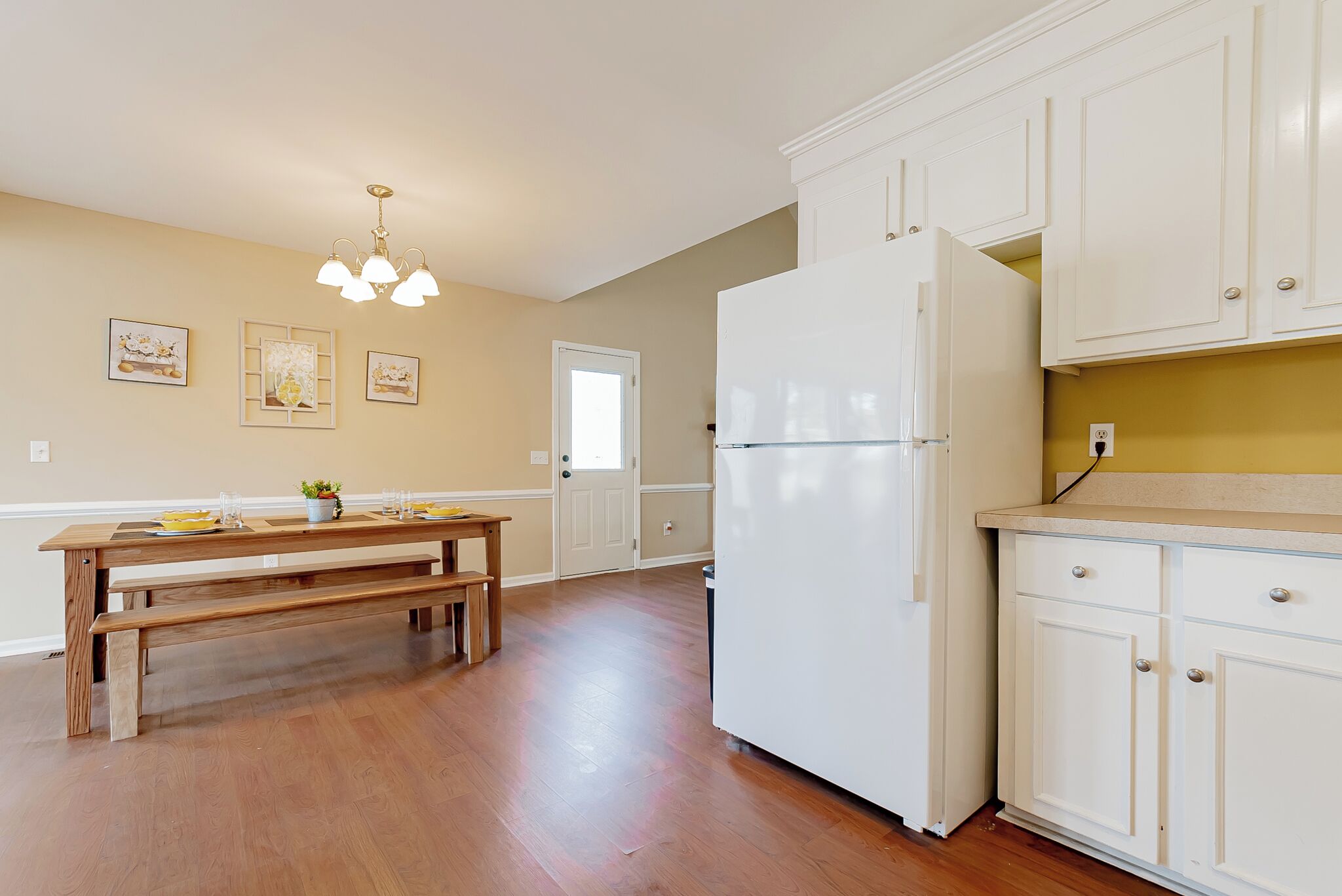 A kitchen with wooden flooring, white cabinets, a refrigerator, and a dining table with benches.