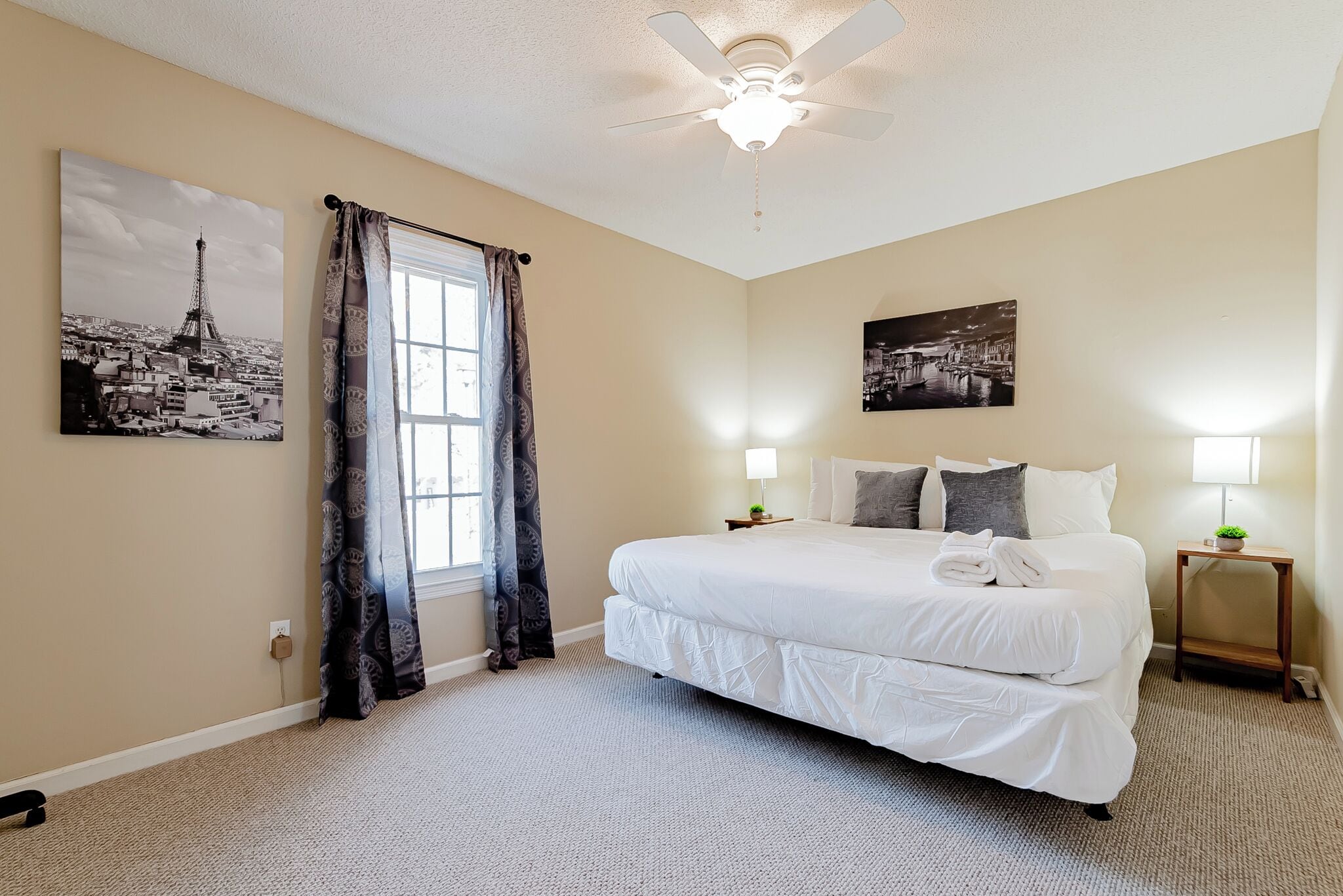 Hotelkamer met een groot bed, twee bedlampen, een plafondventilator en twee kunstwerken aan de muur die stadsgezichten voorstellen.