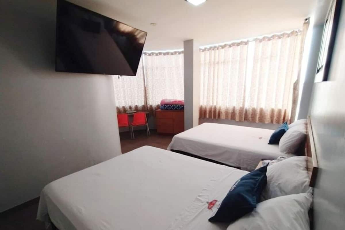 A hotel room with two single beds, a TV mounted on the wall, a small desk with a chair, and a window with curtains.