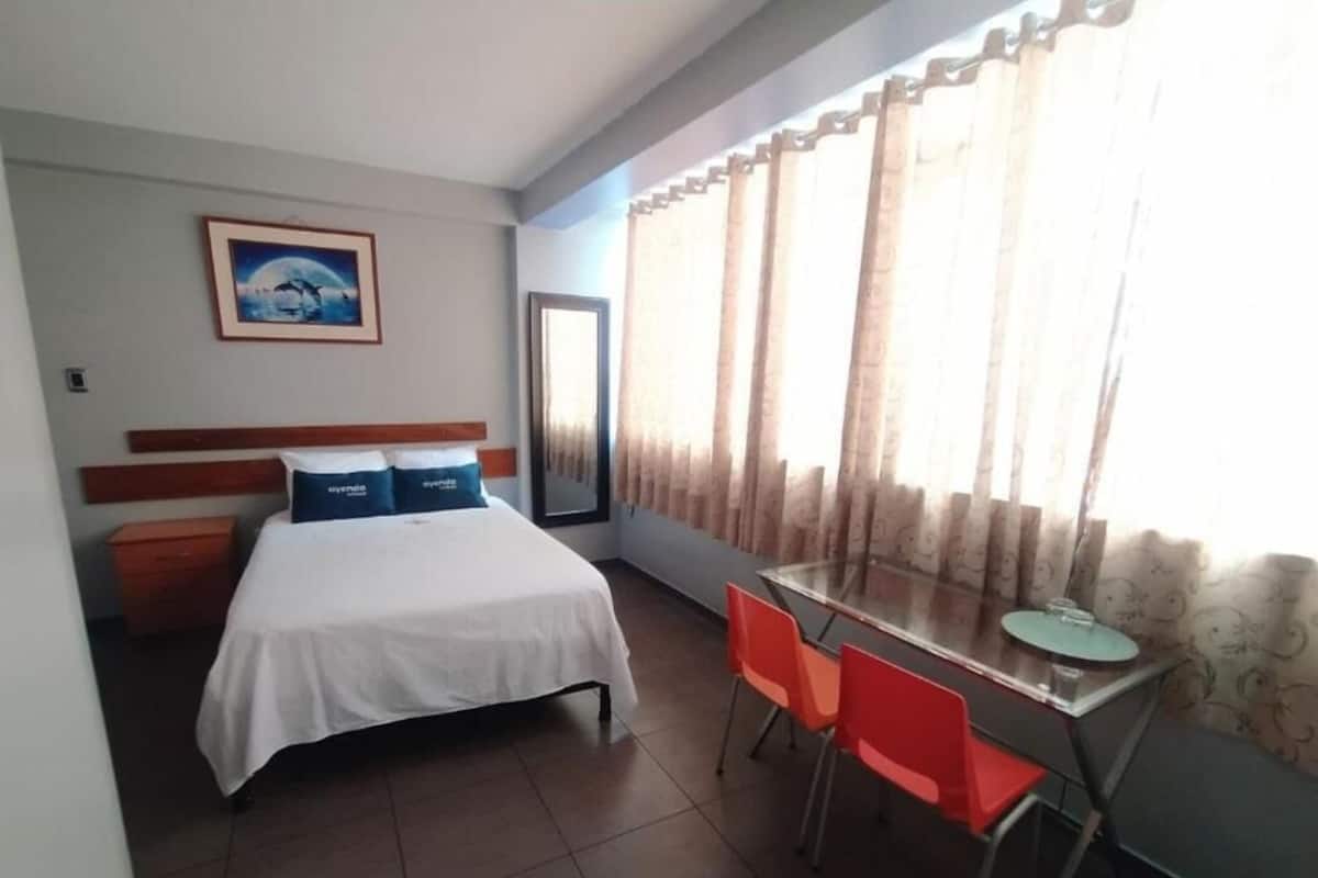 A hotel room with a bed, a desk with two chairs, a large window with curtains, and a framed picture on the wall.