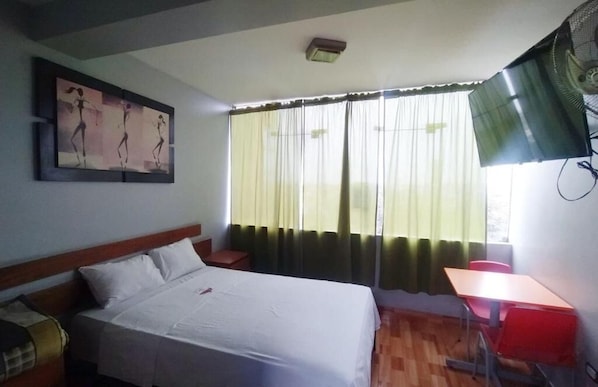 Ett hotellrum med en säng, ett skrivbord, en stol, ett fönster med gardiner och en takfläkt.